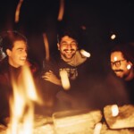 Three friends enjoying a warm campfire at night, sharing smiles and laughter.